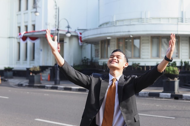 young asian businessman happy and raising hands while standing outdoors with office building