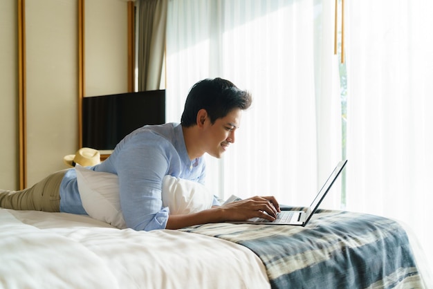 Young Asian businessman lying in bed and working with laptop in room at resort near sea during a summer vacation holiday travel.