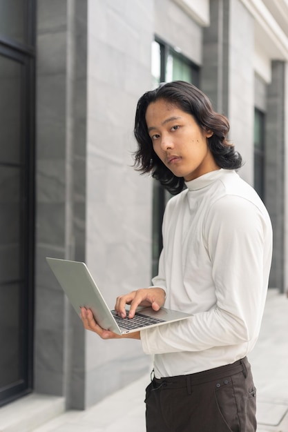 Young Asian businessman using a laptop nearby his office workspace against contemporary corporate buildings in the city