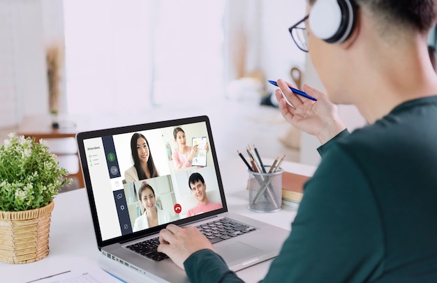 Young Asian businessman wear headphones working remotely from home and virtual video conference meeting with colleagues business people. social distancing at home office concept.