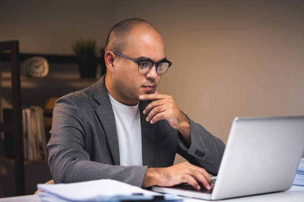 Young asian businessman working with laptop computer in the dark office at night Attractive Indian man work hard overtime serious thinking in home with floor lamp ambient warm light late at night