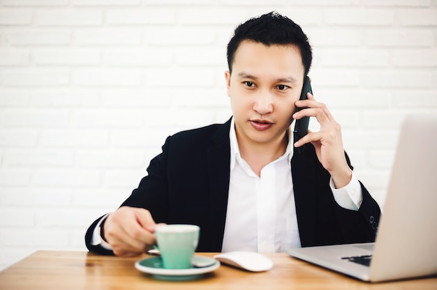 Young asian businessman working with laptop in the modern office, talking on the phone with his hand still holding a cup of coffee.