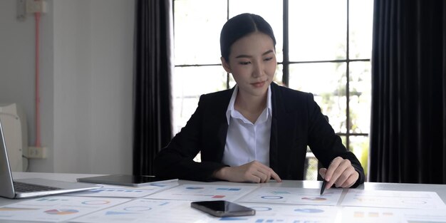 Young Asian businesswoman analyzing report pointing at graph holding a pen using a tablet laptop placed at the office