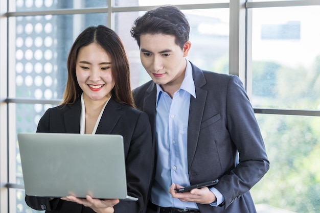 Young Asian businesswoman and businessman partners while working together with laptop computer and business man holding a tablet in office backgroundWorking couple