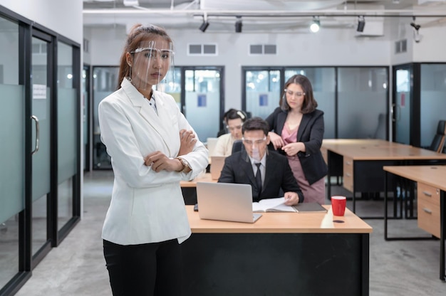 Young asian businesswoman confident with crossed arms wearing face shield and coworkers seriously working on desk at new normal office