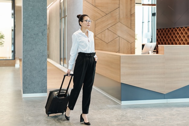 Young Asian businesswoman in formalwear pulling suitcase while waiting for receptionist in hotel lounge