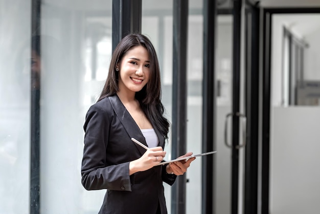 Young Asian businesswoman looking at camera holding pen with tablet at office.