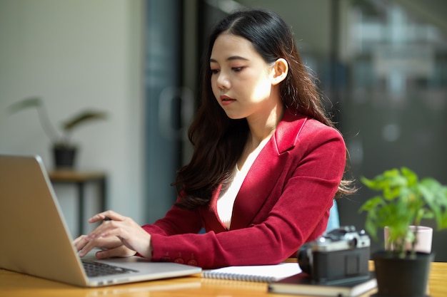 Young asian businesswoman sits at her desk in a  bright modern office workspace working on laptop