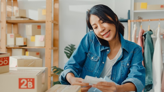 Young asian businesswoman using smartphone receiving purchase order and checking product on stock work at home office