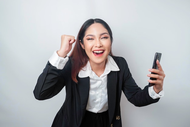 A young Asian businesswoman with a happy successful expression wearing black suit and holding smartphone isolated by white background