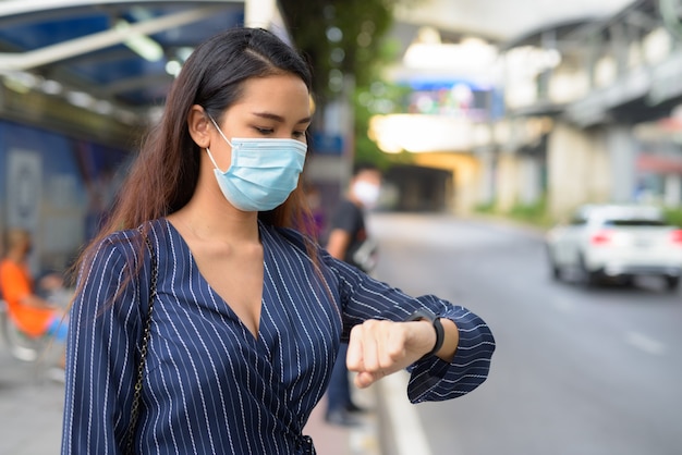 Young Asian businesswoman with mask checking smartwatch and waiting at the bus stop