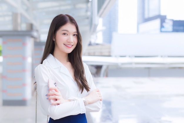 Young Asian confident working woman who has a long hair with a white shirt is arm crossing in hand holding a smartphone standing urban outdoors while walking to office in big city with business buildi