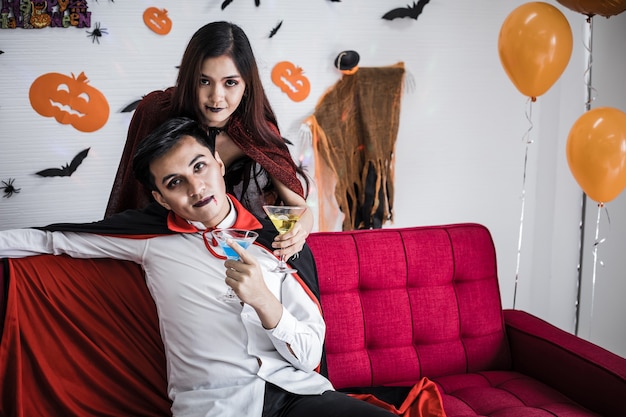 Young Asian couple in costume witch and dracula with celebrate Halloween party and drink wine together in Halloween festival on red sofa in the room at home.