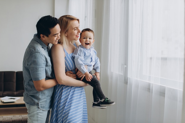 Young asian dad and caucasian blonde mom hold son in her arms in bright room. Multicultural trendy concept