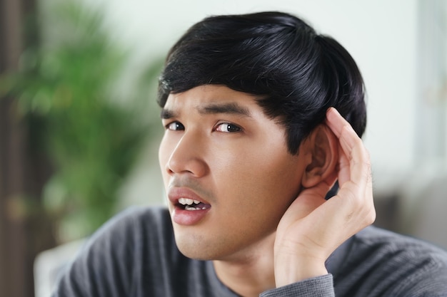 Young Asian deaf disabled man having hearing problems holds his hand over the ear, listens carefully, hard of hearing.