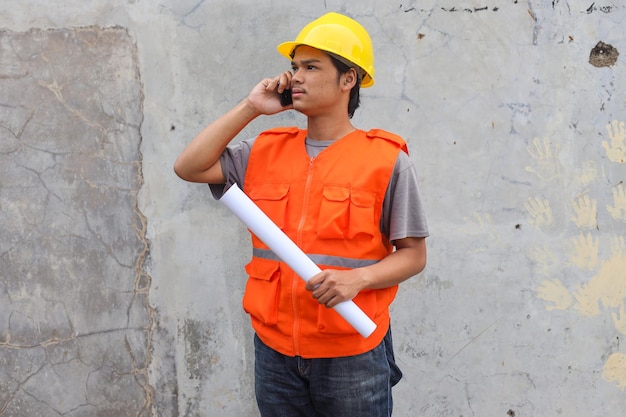 Young Asian engineer in yellow safety helmet using mobile phone to make a call while holds blueprint