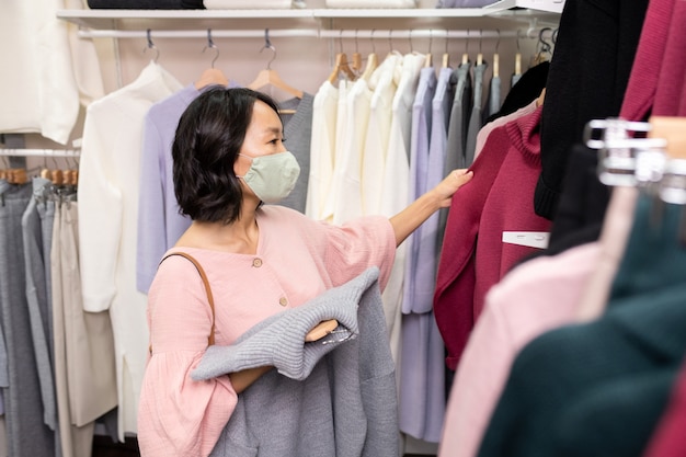 Young Asian female customer in mask and pink smart blouse standing by rack with new assortment of casualwear and choosing sweater