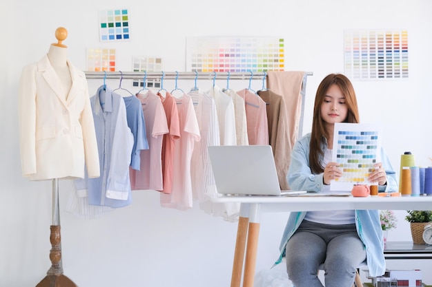 Young Asian female fashion designer choosing color from color samples chart in design studio