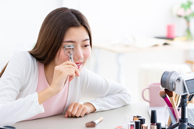 Photo young asian girl beauty vlogger testing new eyelash curler and recording video with camera at home