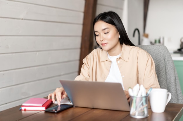 Young asian girl checking smartphone app while working on laptop using devices while doing homework ...