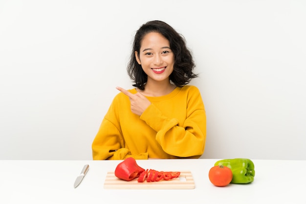 Young asian girl with vegetables in a table pointing finger to the side