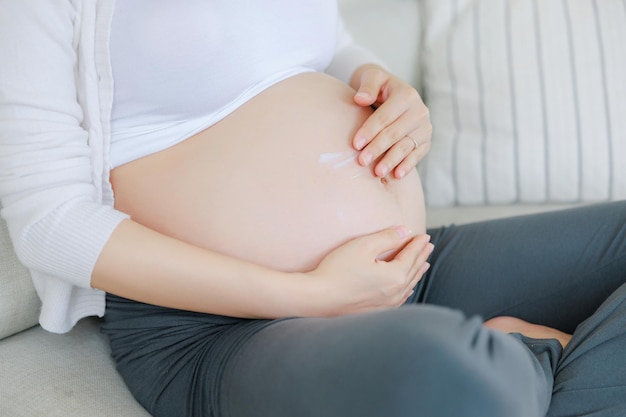 Young Asian happy pregnant woman is sitting on bed and apply cream or serum on belly Family plan pregnancy motherhood people and expectation child and Maternity prenatal care concept