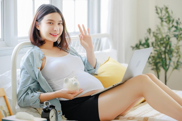 Young Asian happy pregnant woman is sitting and relaxing on bed and touching her belly Family plan pregnancy motherhood people and expectation concept