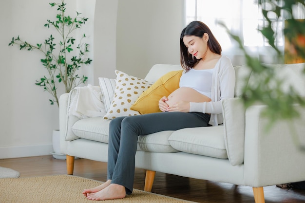 Young Asian happy pregnant woman is sitting on sofa and touching her belly Family plan pregnancy motherhood people and expectation Child and Maternity prenatal care concept