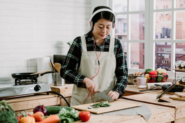 Young asian housewife in headphones and apron listening music and enjoying cooking preparation in modern kitchen at home. Clean healthy food and diet lifestyle concept. female cutting cucumber slice