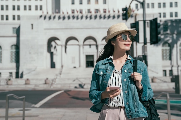 young asian local woman hold mobile phone with backpack looking smiling enjoy sunshine on face walking in Los Angeles Downtown City Hall California USA. beautiful girl with cellphone relax outdoor