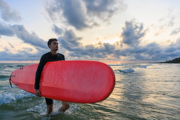 Young Asian male surfer holding a board and walking back to shore after an extreme water sports practice