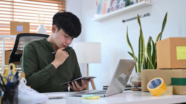 Young Asian man checking online order on digital tablet and preparing parcel boxes for delivery to customers