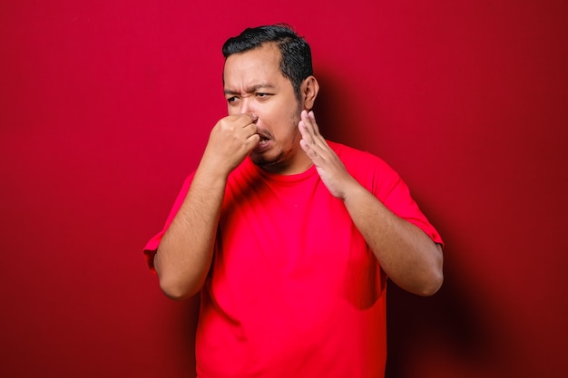 Young Asian man close his nose for bad smell, isolated on red background