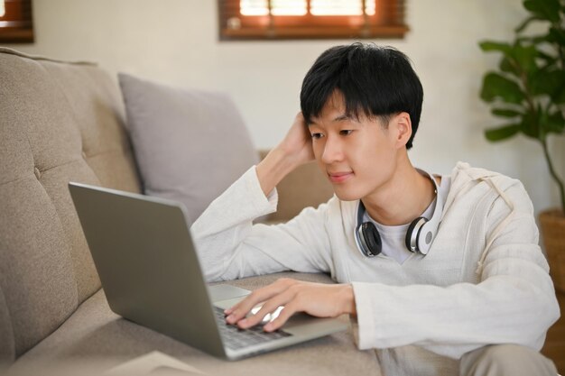 Young Asian man in comfy clothes using laptop on a sofa in his living room