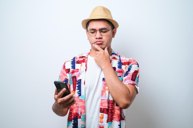 Young asian man holding a phone looking sideways with doubtful and skeptical expression