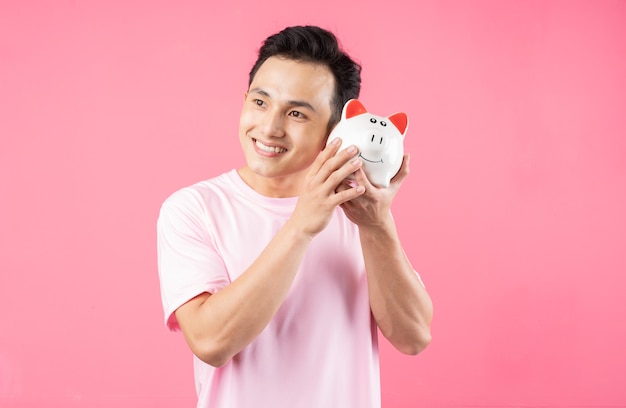 Young Asian man holding piggy bank on pink