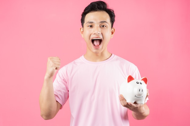 Young Asian man holding piggy bank on pink