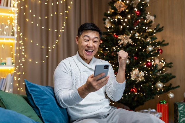 A young asian man is playing games on the phone sitting at home on the couch near the nineyearold