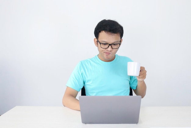 Young Asian man is serious when work in laptop with drink coffee in white glass Indonesia Man wear blue shirt Isolated grey background