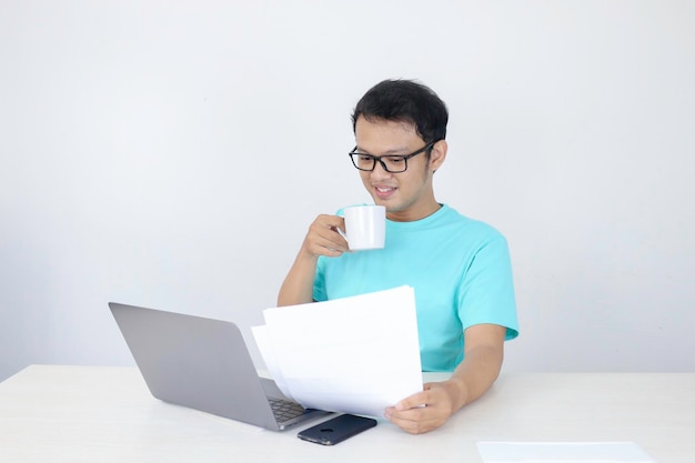 Young Asian Man is smile and happy when working on a laptop and document with white mug on hand Indonesian man wearing blue shirt