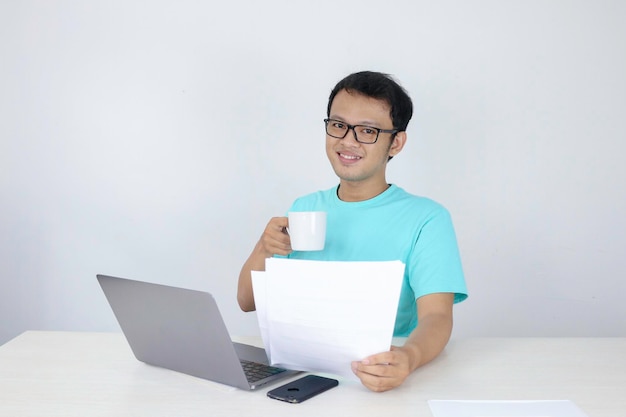 Young Asian Man is smile and happy when working on a laptop and document with white mug on hand Indonesian man wearing blue shirt