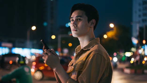 Young Asian man is using his phone while walking in the street at night