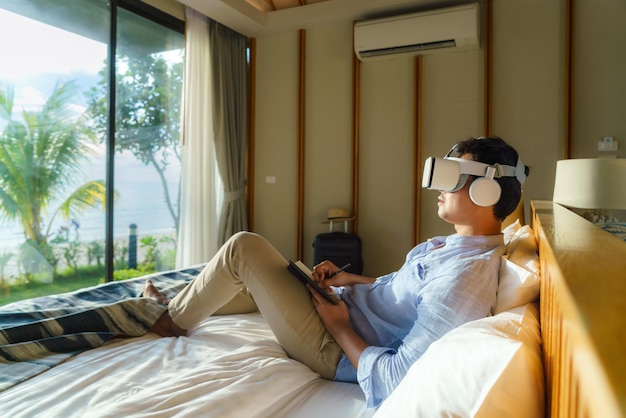 Young Asian man lying on bed and using virtual reality glasses for business meeting during his travel summer vacation holiday.