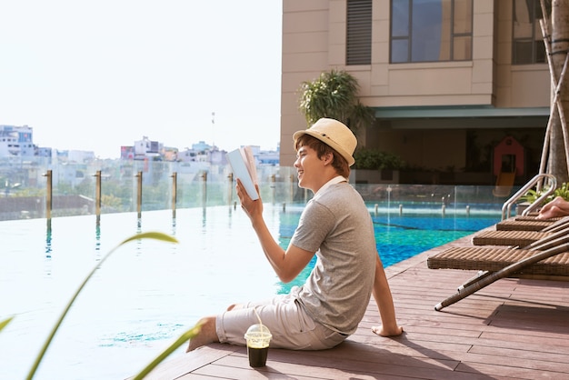 Young asian man reading book by the pool on a sunny summer day