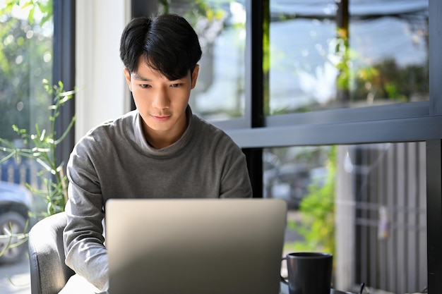 A young Asian man remote working at the coffee shop focusing on his work on laptop