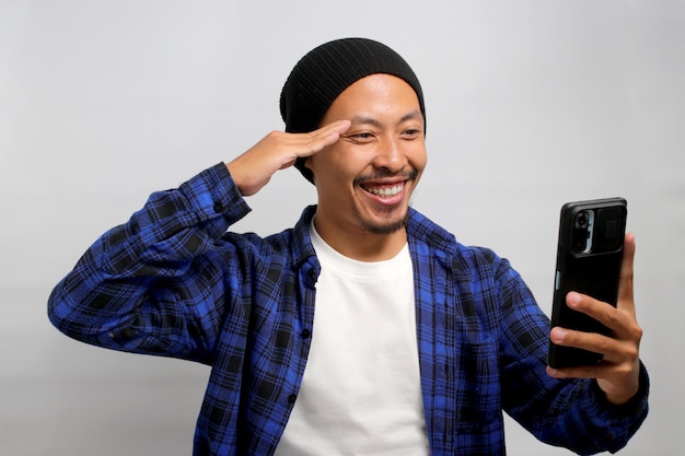 A young Asian man salutes while taking a selfie with his smartphone against a white background