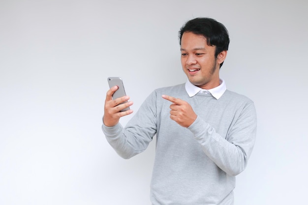 Young Asian man shocked and happy with what he see in the smartphone on isolated grey background