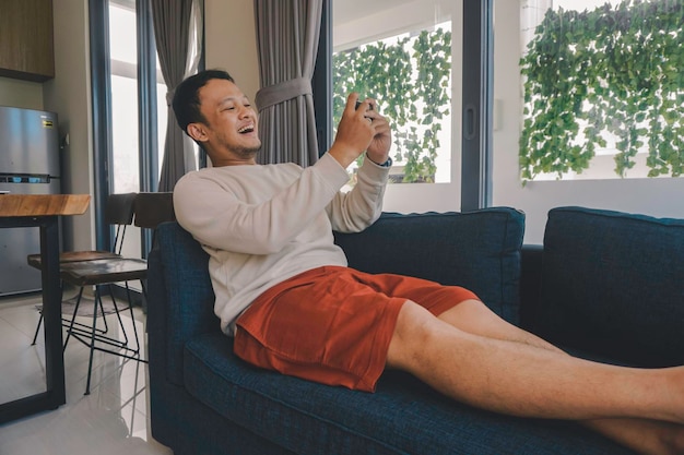 Young Asian man spend his free time at home sitting on a cozy sofa in the living room texting message on mobile app on his smartphone