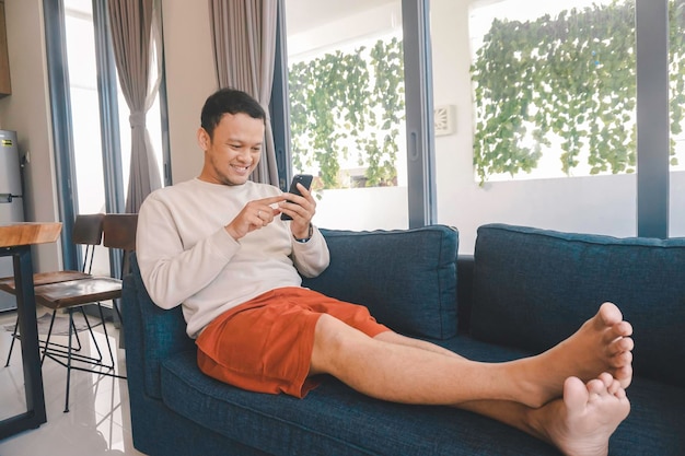 A young Asian man spends his free time at home sitting on a cozy sofa in the living room pointing to his smartphone