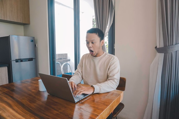 A young Asian man spends his time at home sitting in the dining room shocked while looking at his laptop